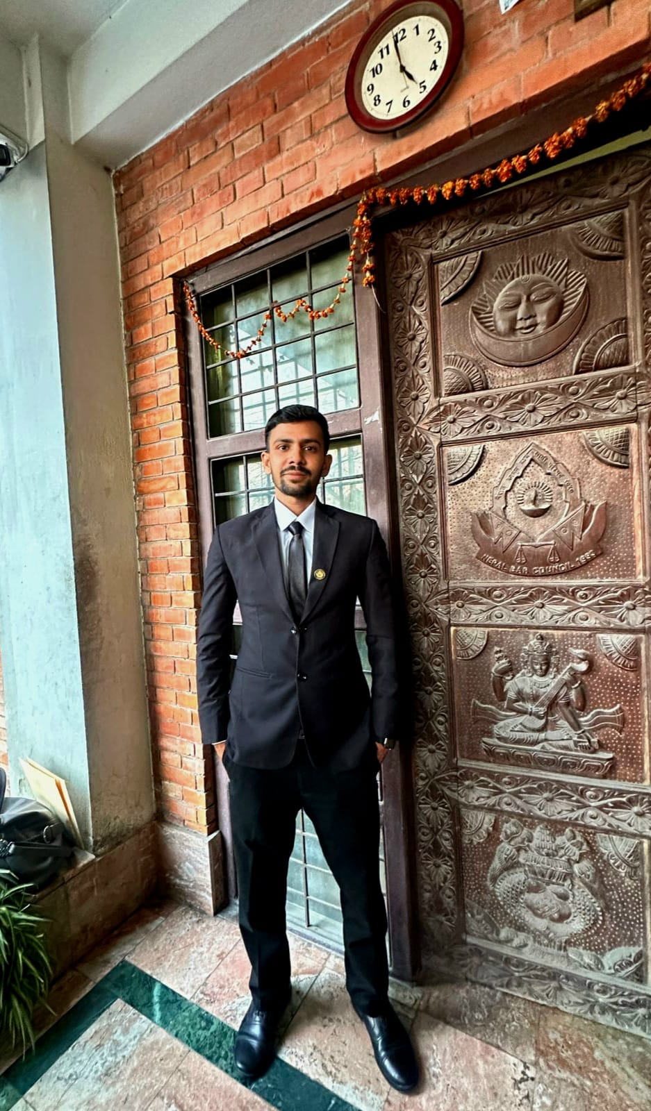 Legal advisor of virohana in a suit standing in front of an ornate wooden door with a clock above it.