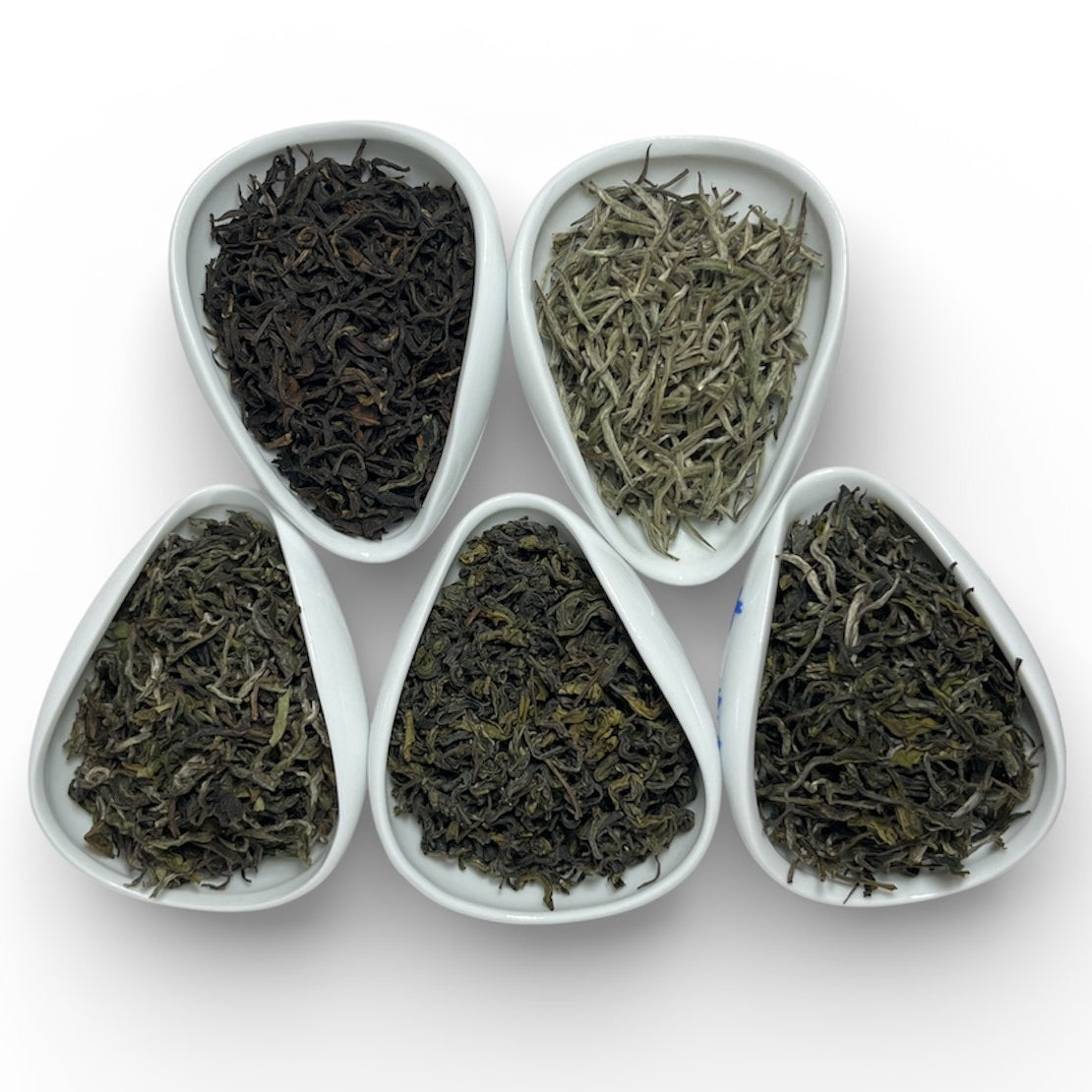 Five white heart-shaped bowls filled with different types of tea leaves on a white background