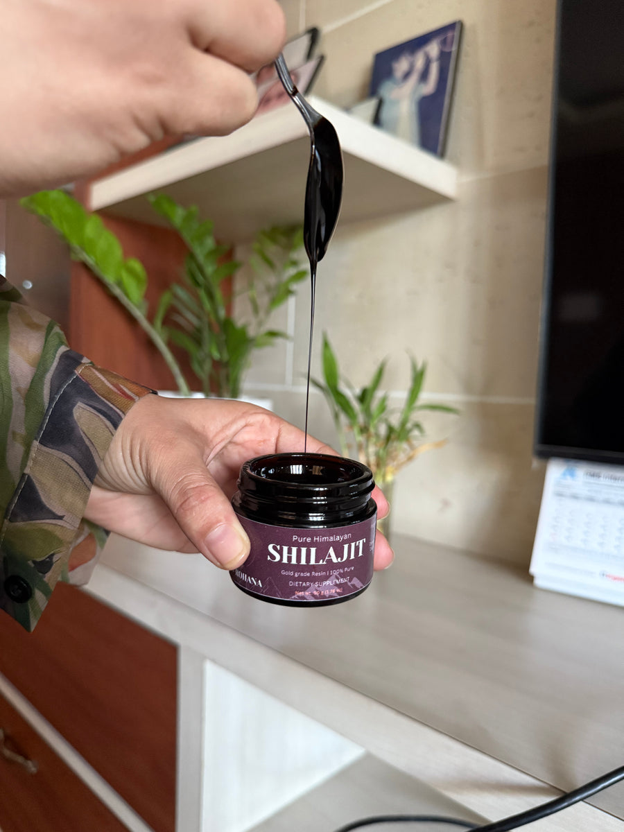 Person using a spoon to scoop out a dark substance from a purple jar labeled 'SHIL-JIT' on a kitchen counter.