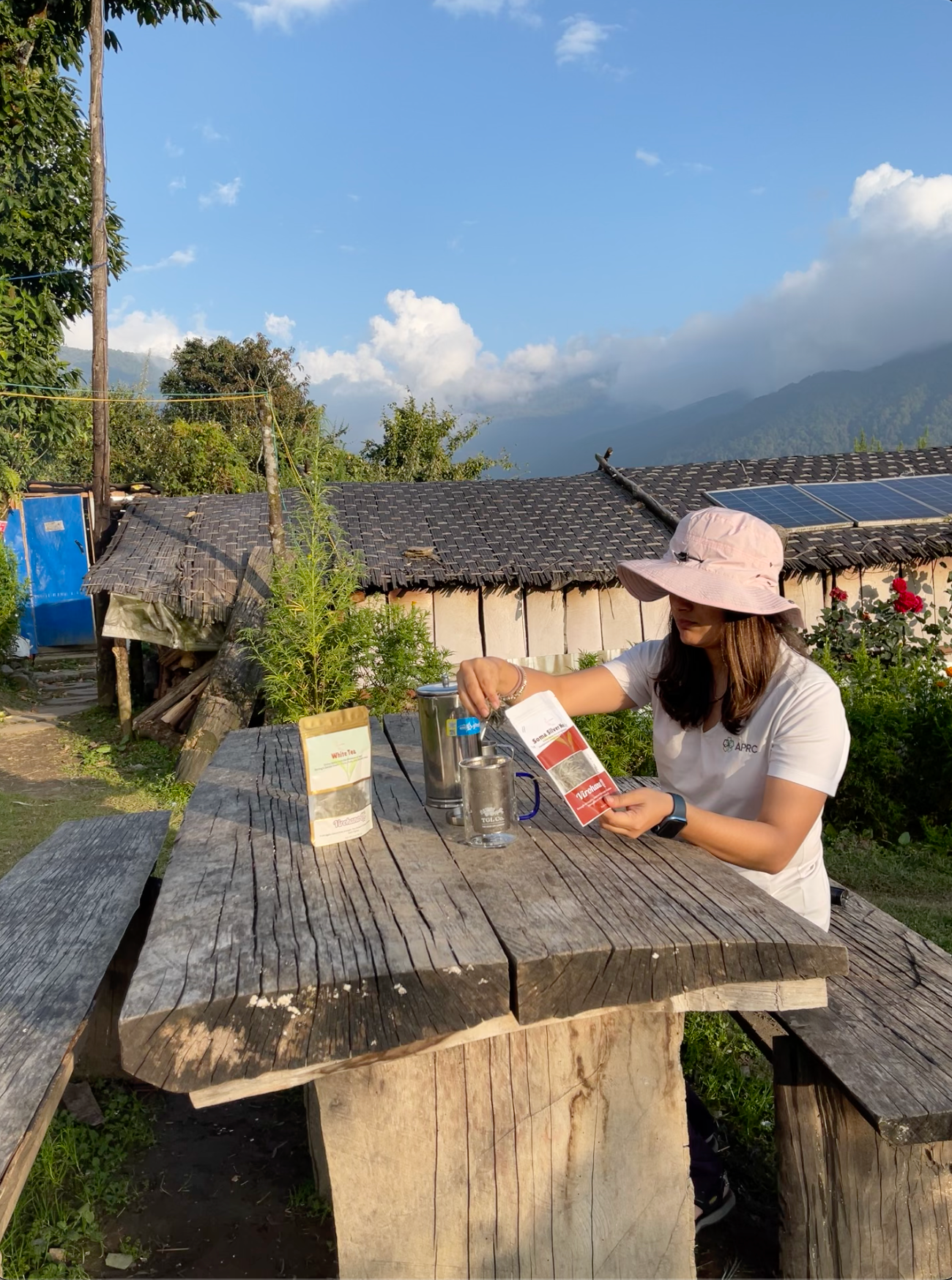 Person sitting at a wooden table outdoors with mountains in the background preparing soma silver needles tea